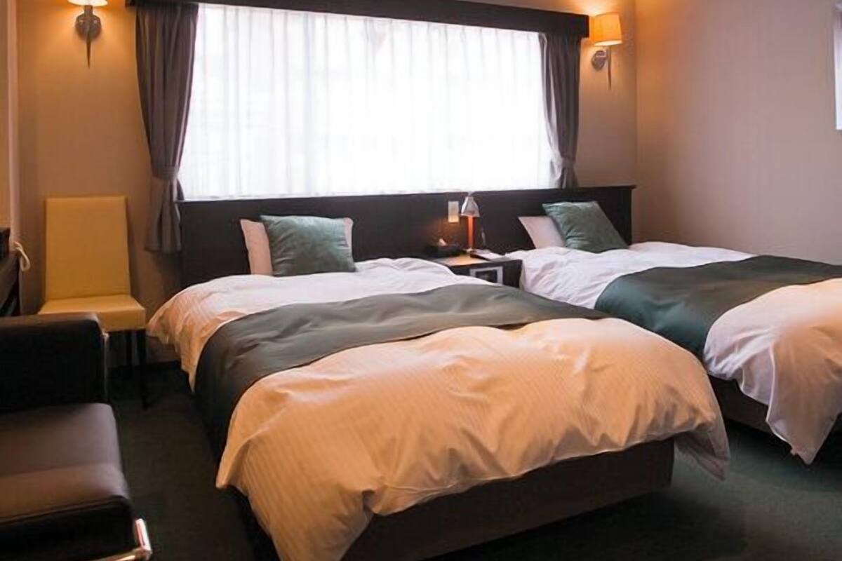 Ein Hotelzimmer mit zwei Betten, einem Sessel, einem Nachttisch und einem Fenster mit Vorhängen.