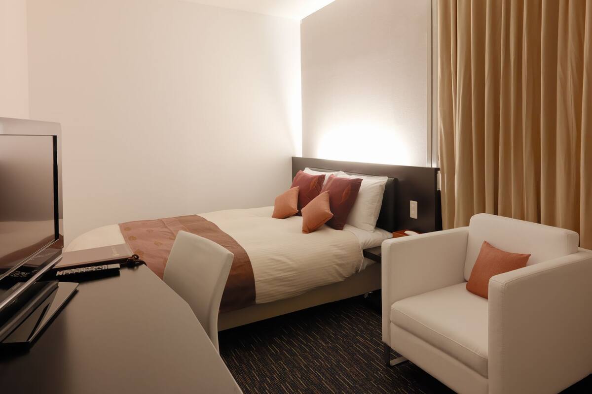 A hotel room with a bed, a desk with a television, a chair, and a curtain.