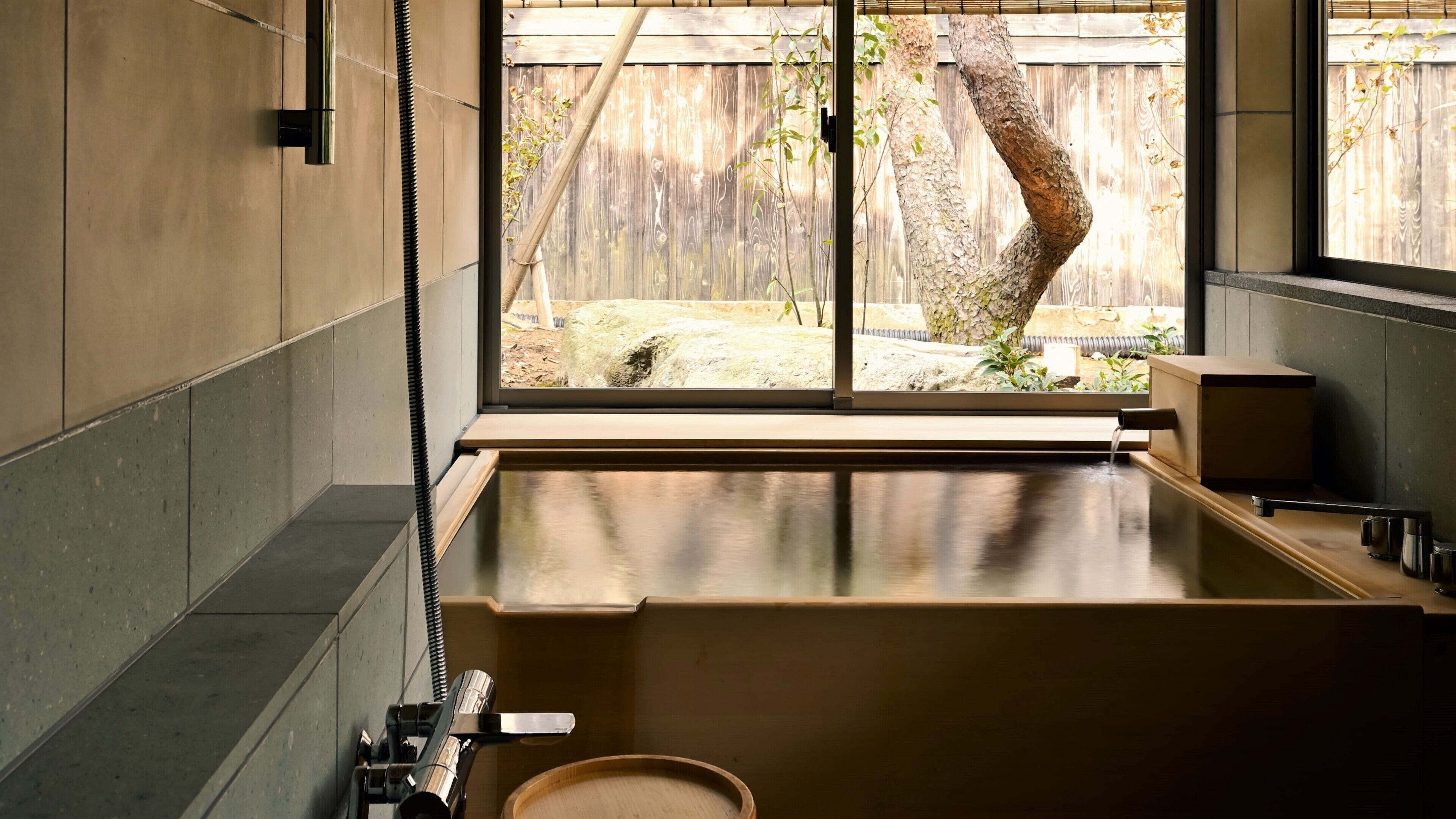 A modern bathroom with a large bathtub, a window with a view of a wooden fence and tree, and a bamboo plant outside.
