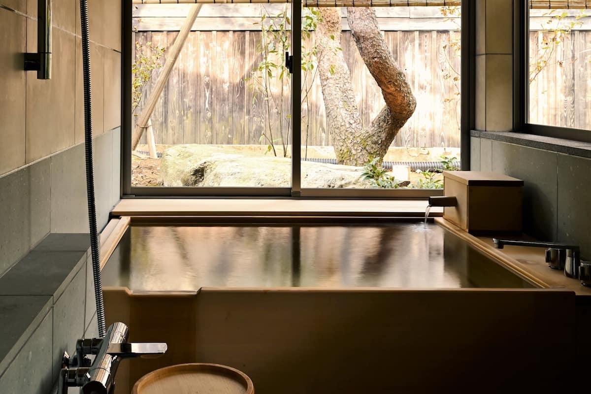 A modern bathroom with a large bathtub, a window with a view of a wooden fence and tree, and a bamboo plant outside.