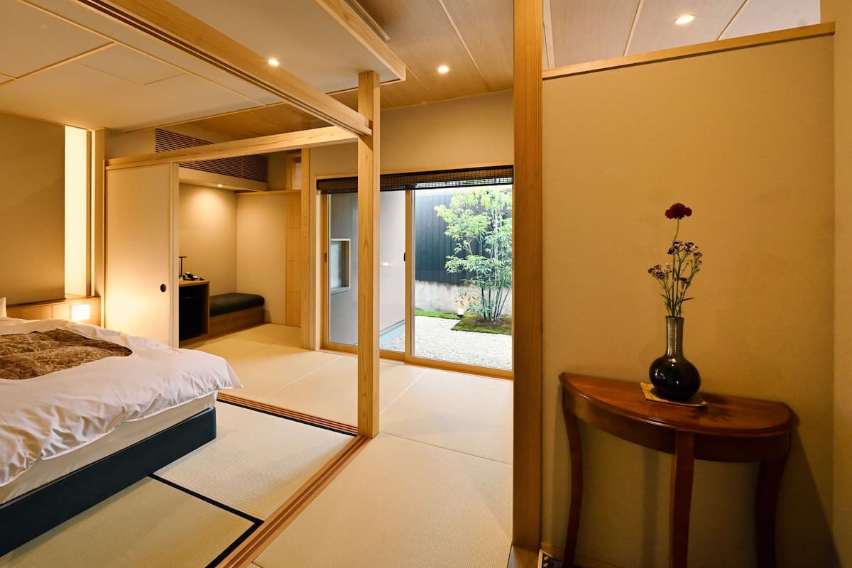 A traditional Japanese-style bedroom with tatami flooring, a bed with white bedding, a small table with a vase, and a sliding door leading to a garden.