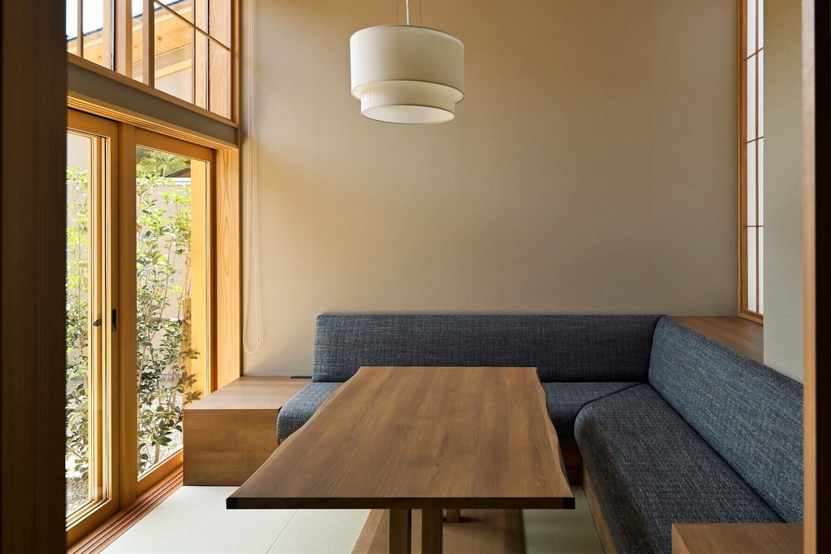 A room with a wooden table, a blue sofa, a hanging light fixture, and a view of greenery through a window.