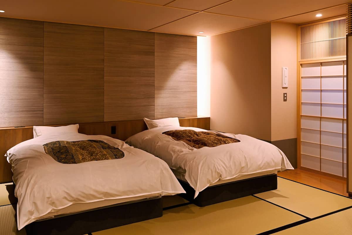 A hotel room with two beds, a wooden headboard, a sliding shoji screen, tatami flooring, and a wall-mounted heater.