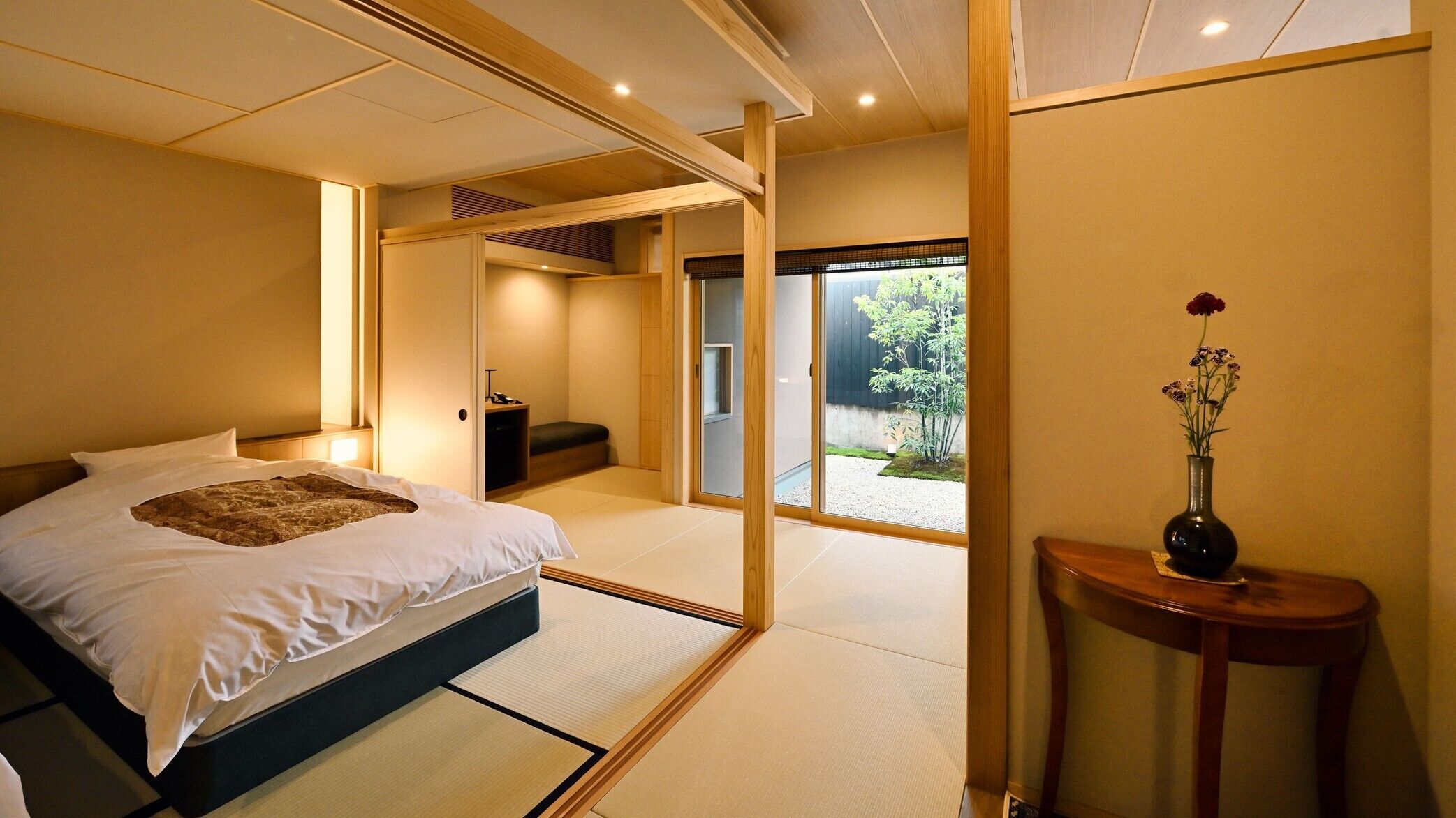 A traditional Japanese-style bedroom with tatami flooring, a bed with a white and brown bedspread, and a vase with flowers on a side table.