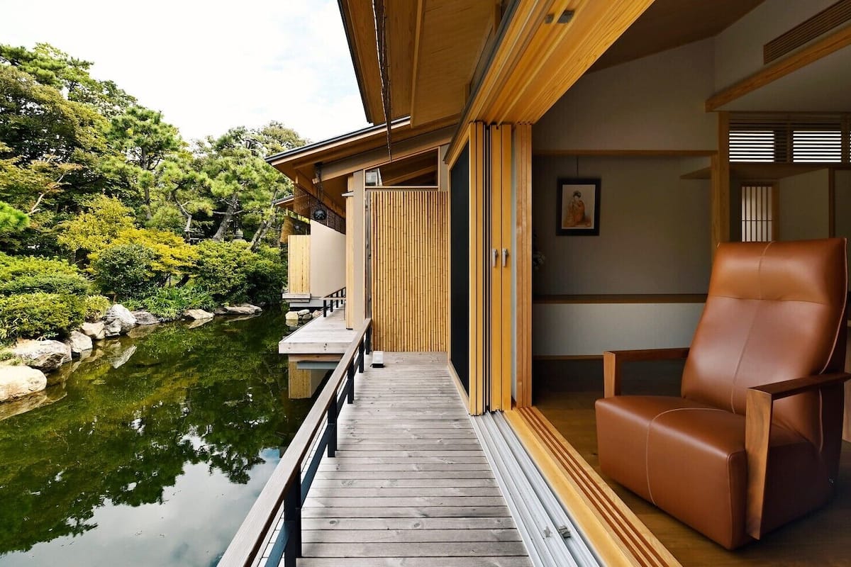 A wooden deck leading to a traditional Japanese-style room with a view of a pond and greenery.