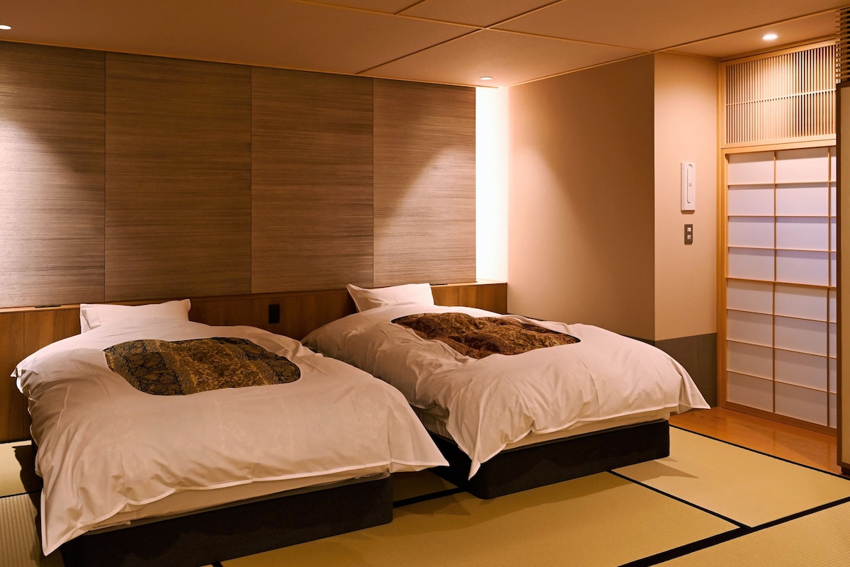 A hotel room with two beds, a wooden headboard, a sliding shoji screen, tatami flooring, and a wall-mounted heater.