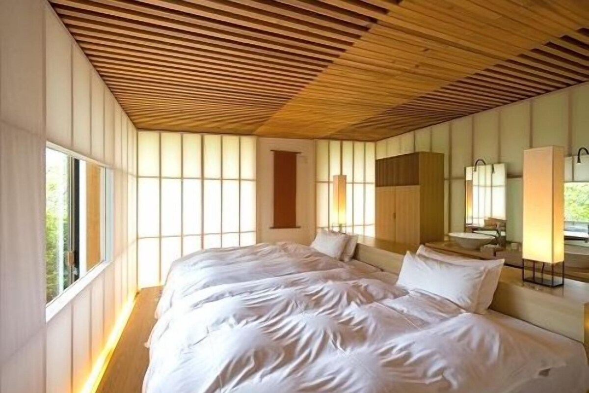 A bedroom with a wooden slatted ceiling, a large bed with white linens, a bedside table, and a mirror.
