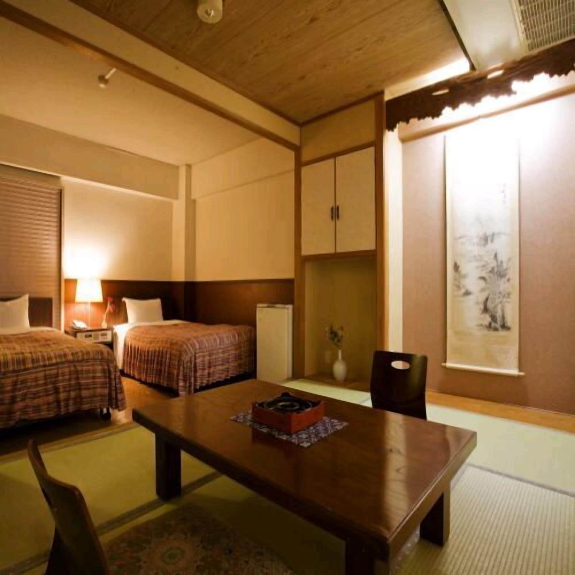 A hotel room with two beds, a wooden table, a chair, a desk, a lamp, a vase, and a painting on the wall.