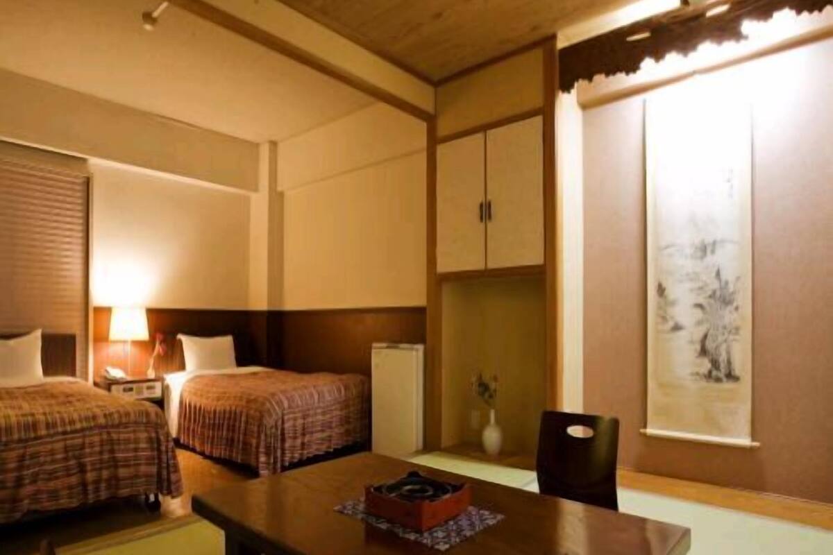 A hotel room with two beds, a wooden table, a chair, a desk, a lamp, a vase, and a painting on the wall.