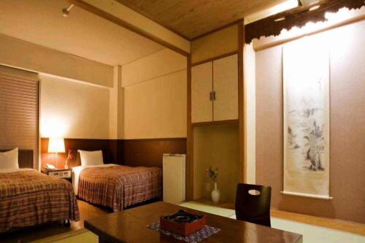 A hotel room with two beds, a wooden table, a chair, a desk, a lamp, a small cabinet, and a window with a view.