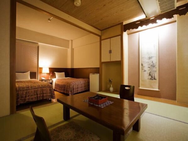 A room with two beds, a wooden table, a chair, a small cabinet, and a window with a view. - Takamatsu Hotel Sakika (Takamatsu)