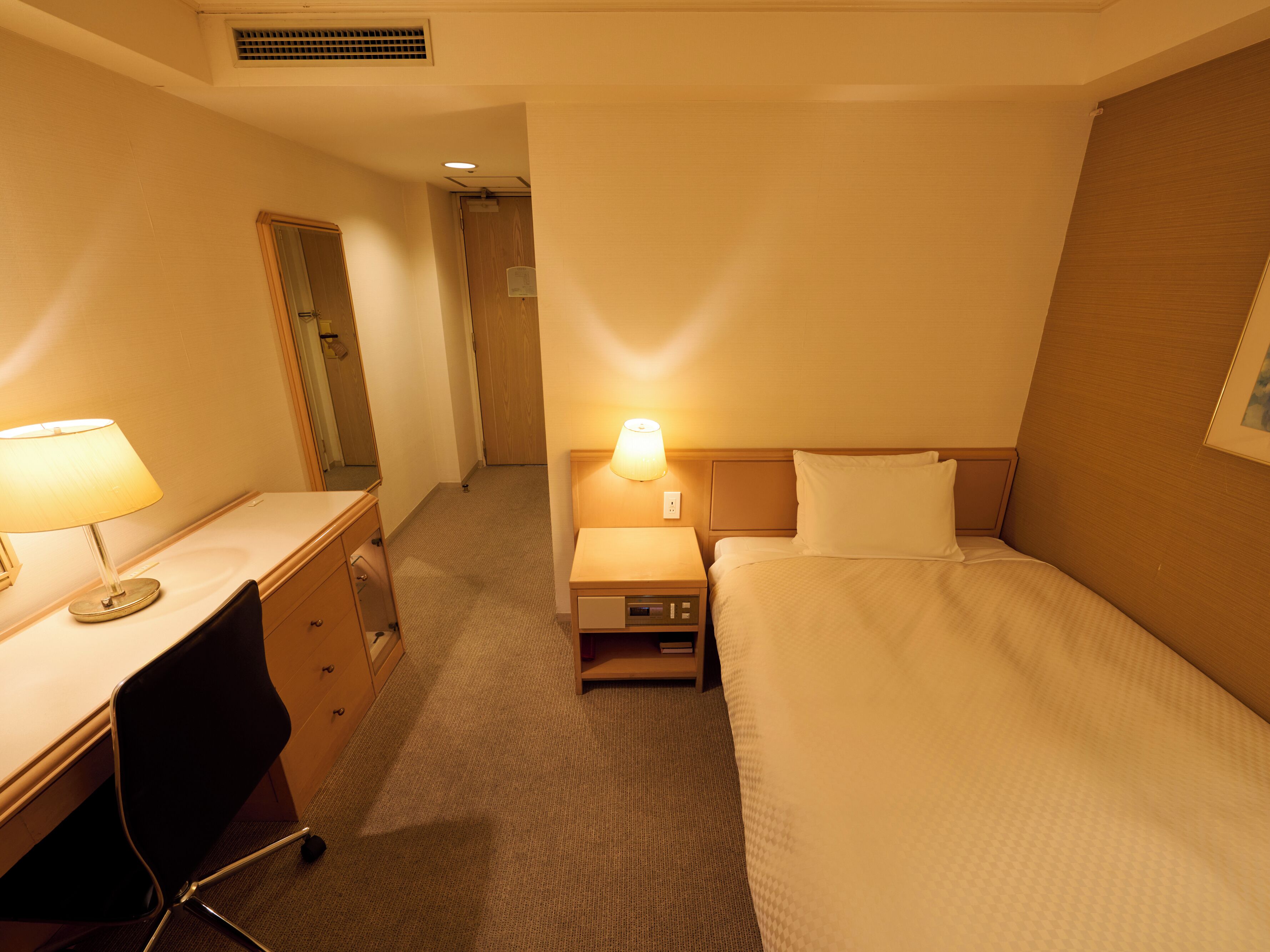 A hotel room with a bed, a desk with a lamp, a chair, a nightstand with a lamp, and a door leading to another room.