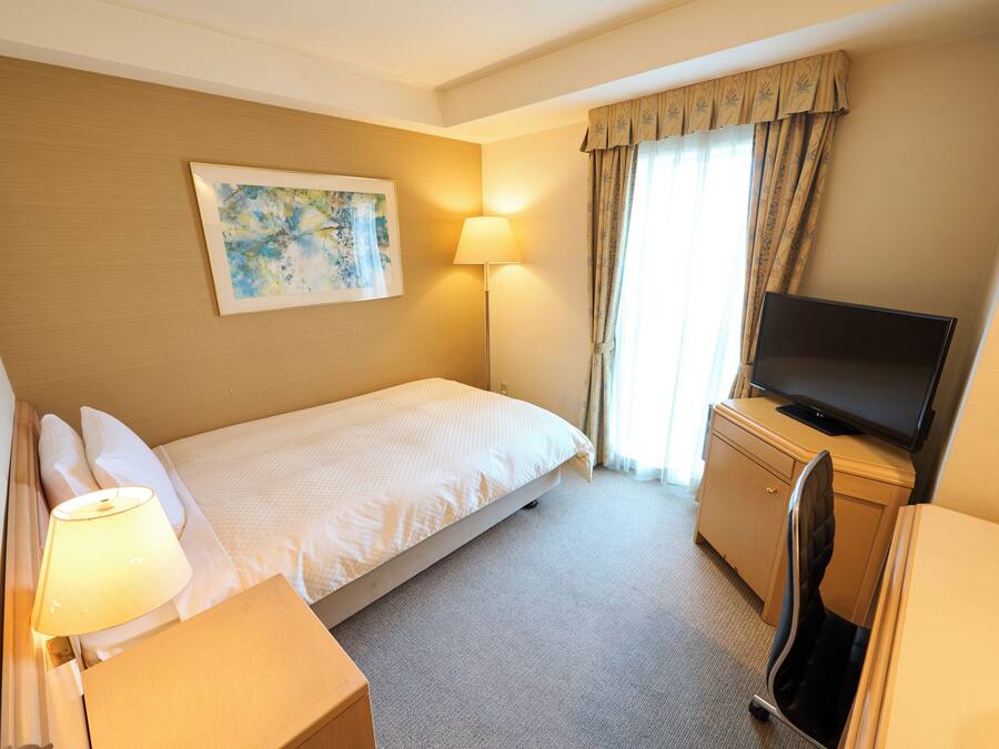 Quarto de hotel com cama, escrivaninha, televisão, candeeiro e um quadro na parede.