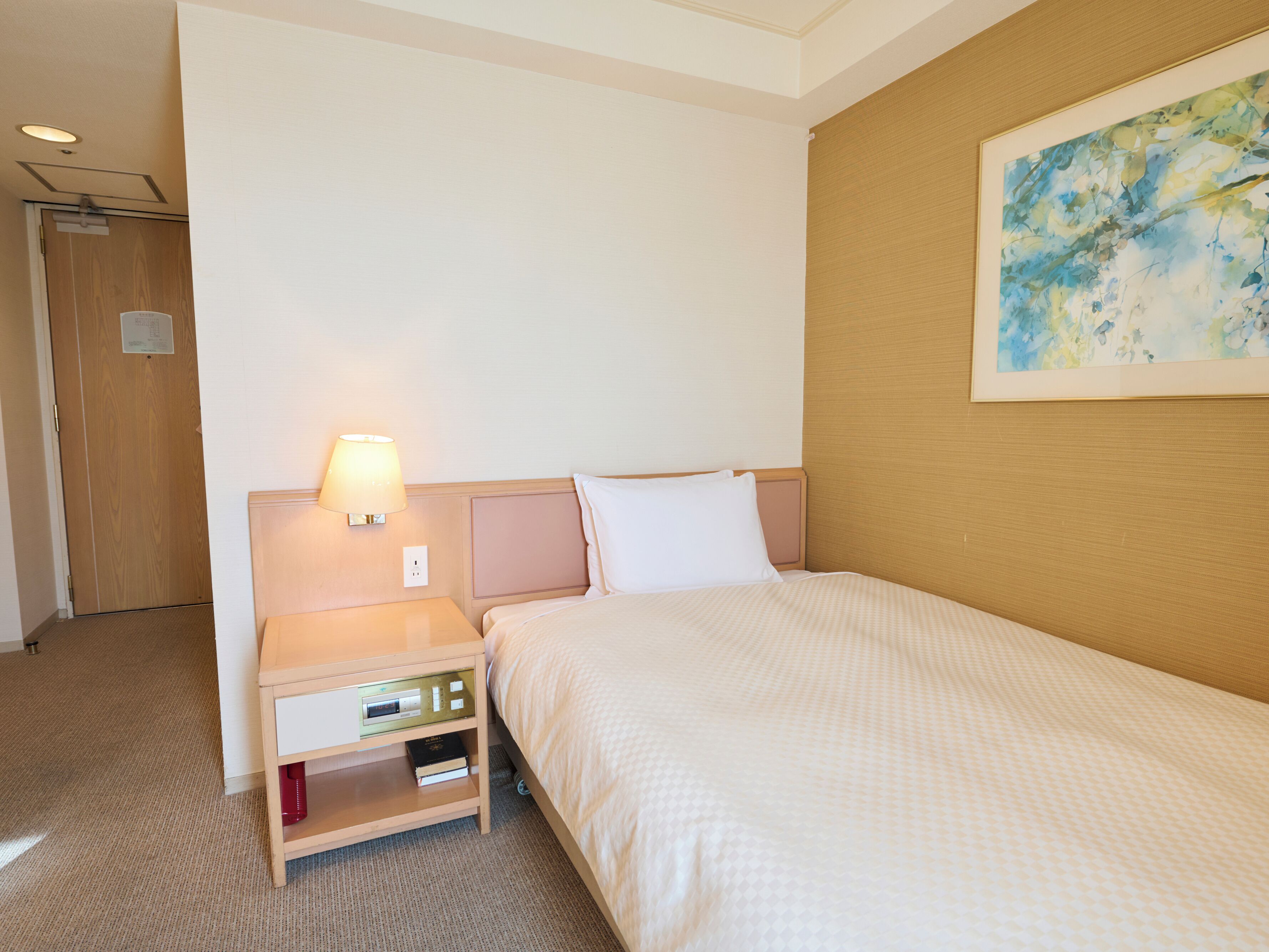 A hotel room with a bed, a nightstand with a lamp, a wall-mounted painting, and a door.