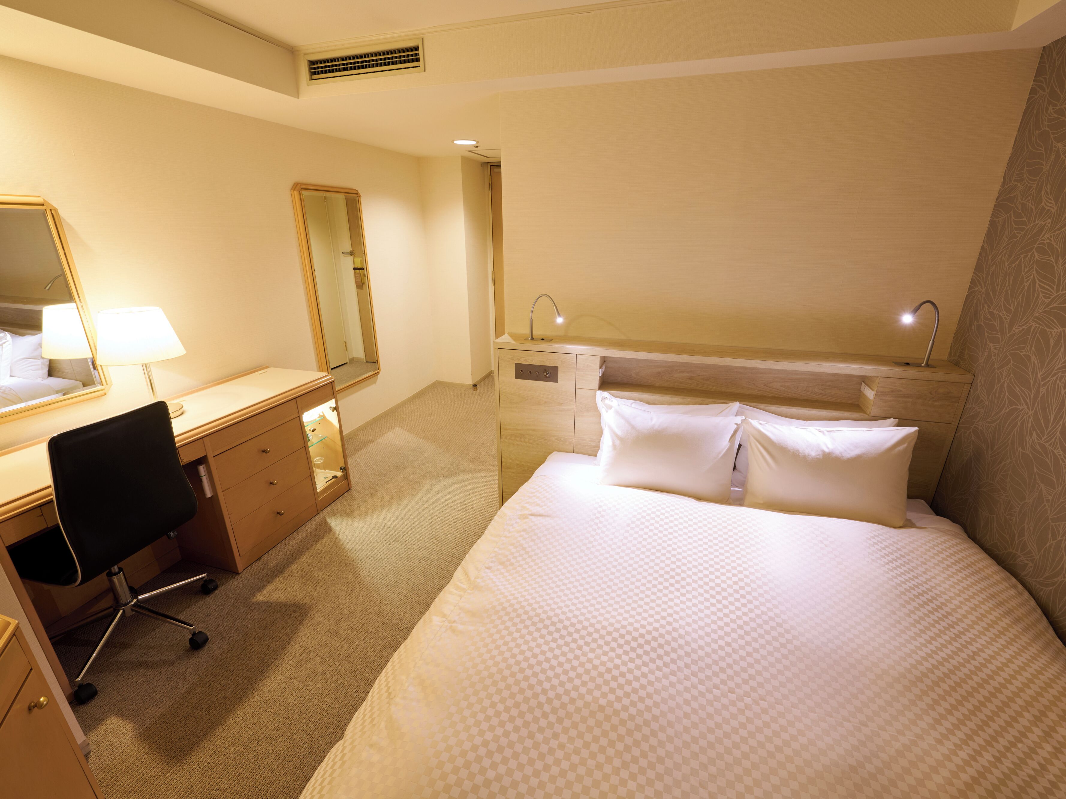 A hotel room with a bed, a desk with a chair, a mirror, a bedside table with a lamp, and a wardrobe.