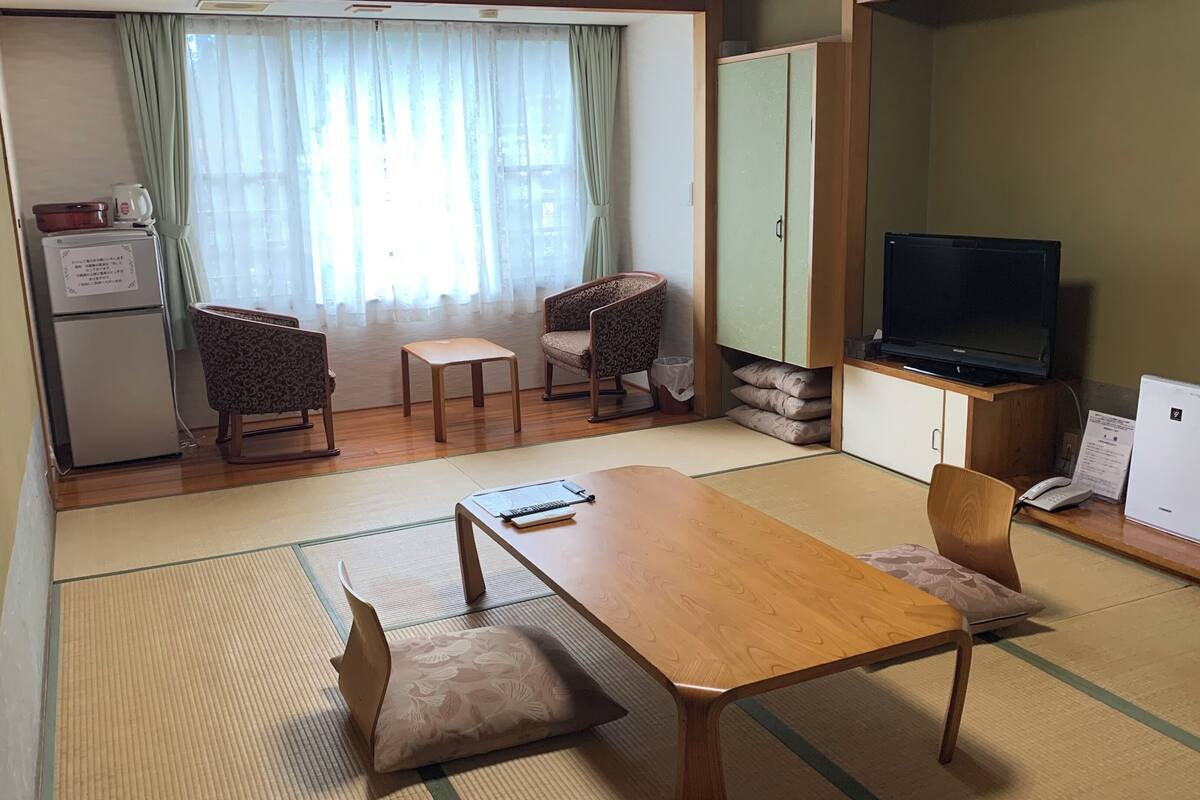 Une pièce de style japonais traditionnel, avec un sol recouvert de tatamis, une table à manger en bois, deux chaises à motifs, une petite table, un téléviseur et un réfrigérateur.