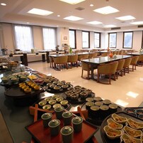 Un comedor amplio con varias mesas y sillas, un bufé con diversos platos, y ventanas grandes que dejan entrar luz natural.