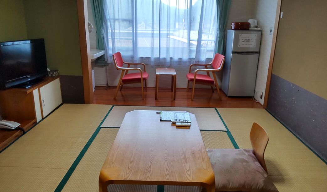 Una habitación con una mesa de madera, dos sillas, un televisor, un refrigerador y una ventana con cortinas.