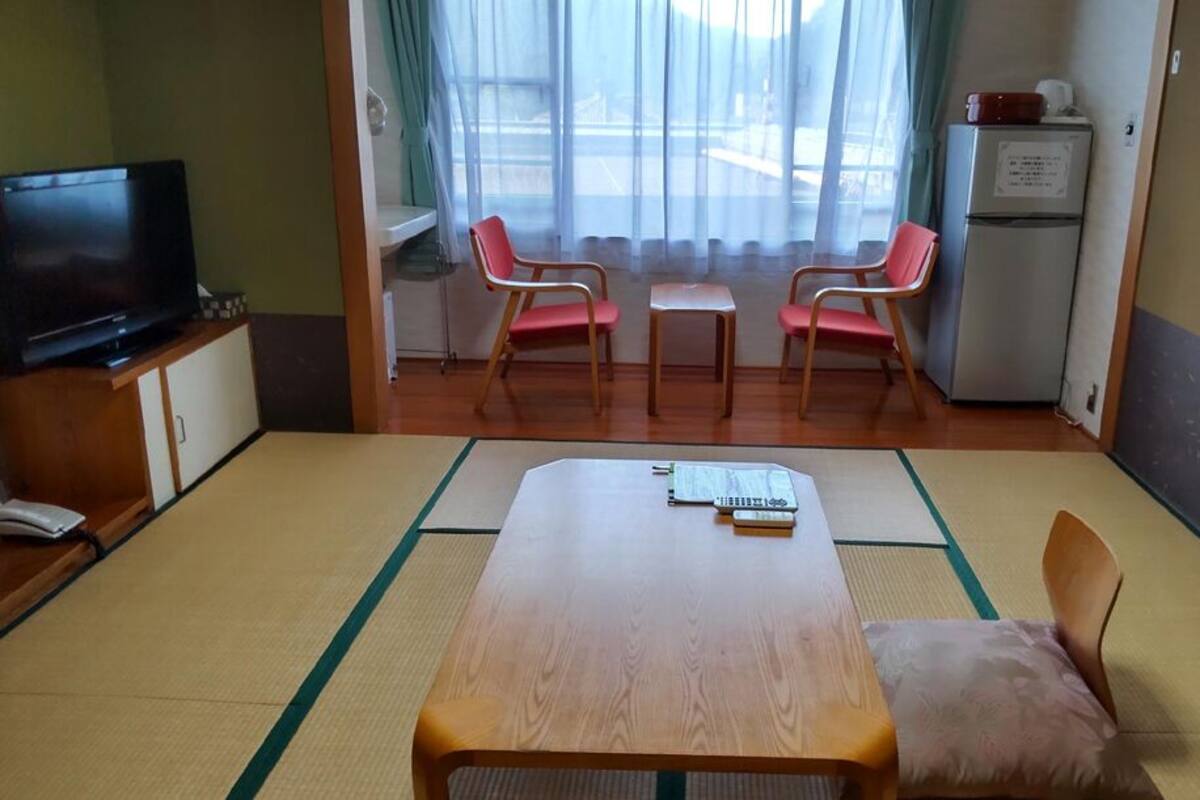 Une pièce avec une table en bois, deux chaises, un téléviseur, un réfrigérateur et une fenêtre avec des rideaux.
