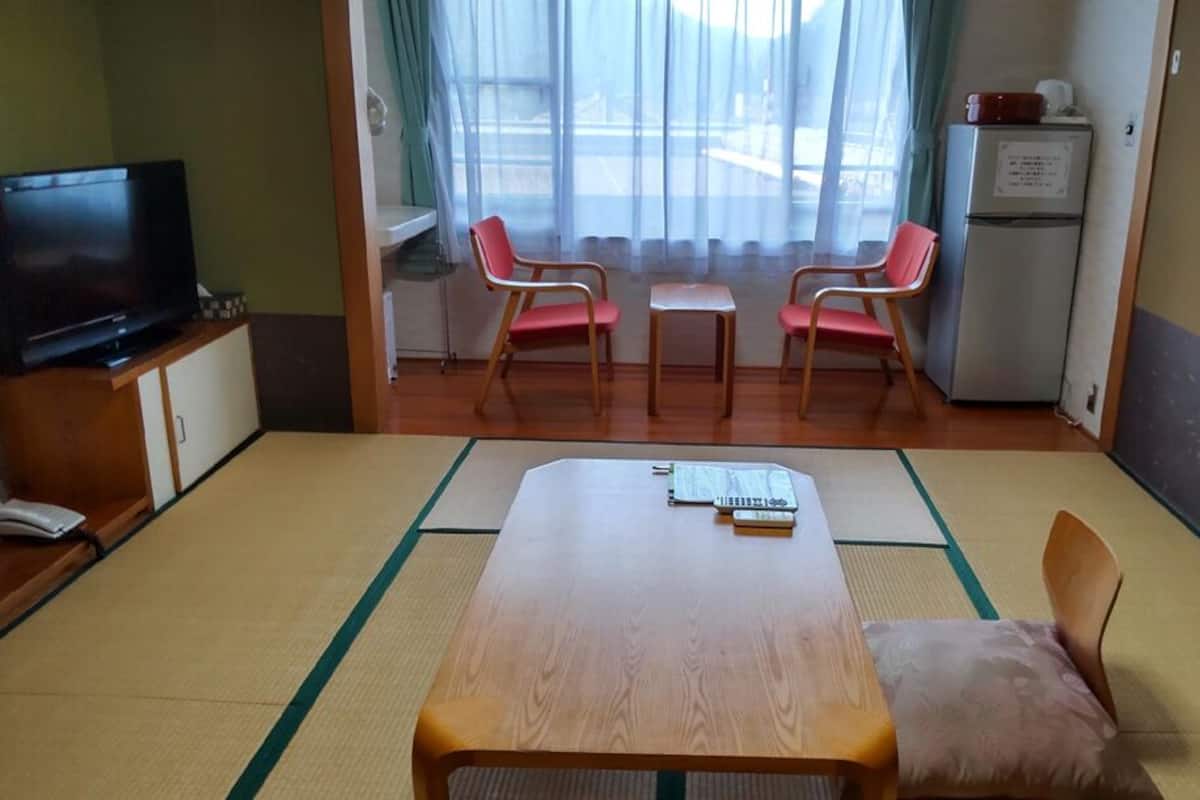 Ein Zimmer mit Holztisch, zwei Stühlen, einem Fernseher, einem Kühlschrank und einem fenster mit Vorhängen.