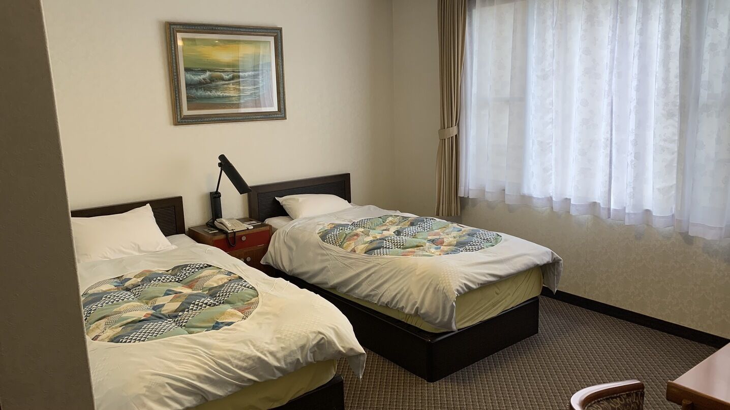 A hotel room with two beds, a painting on the wall, a telephone on the nightstand, and a window with sheer curtains.