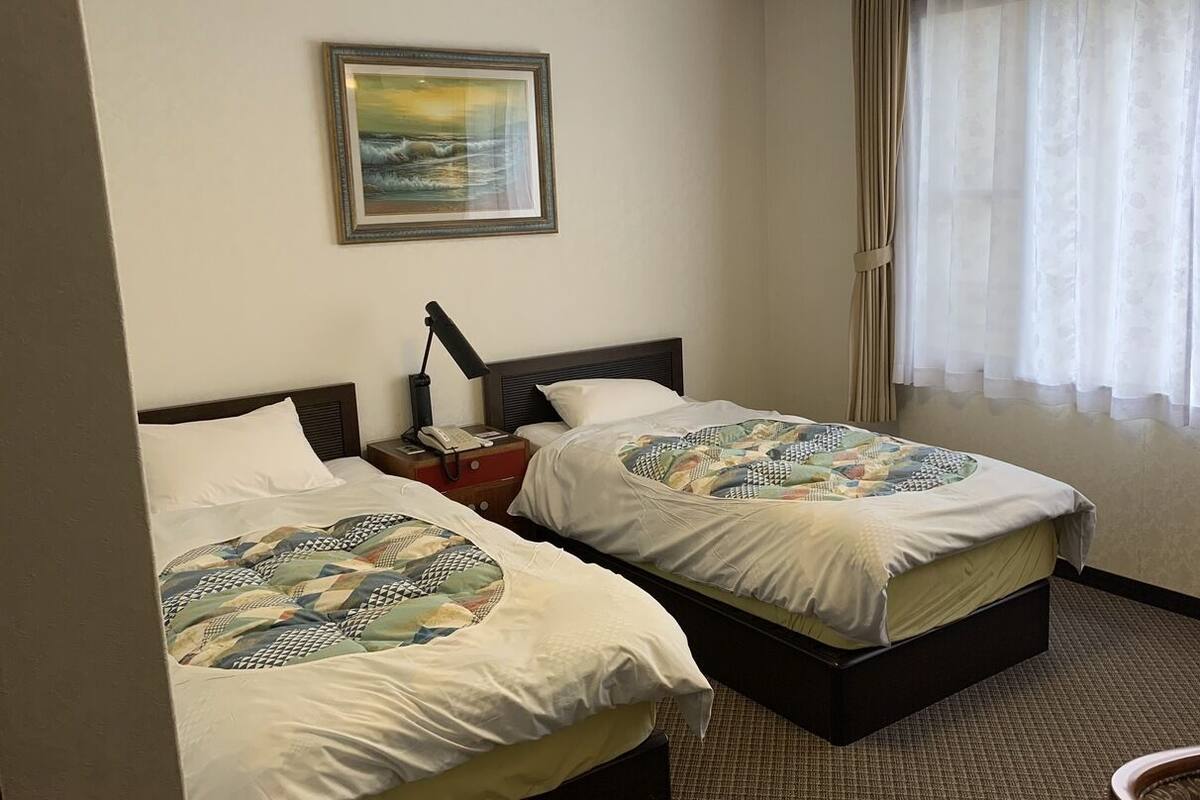 Une chambre d’hôtel avec deux lits, un tableau au mur, un téléphone sur la table de chevet et une fenêtre avec des rideaux légers.
