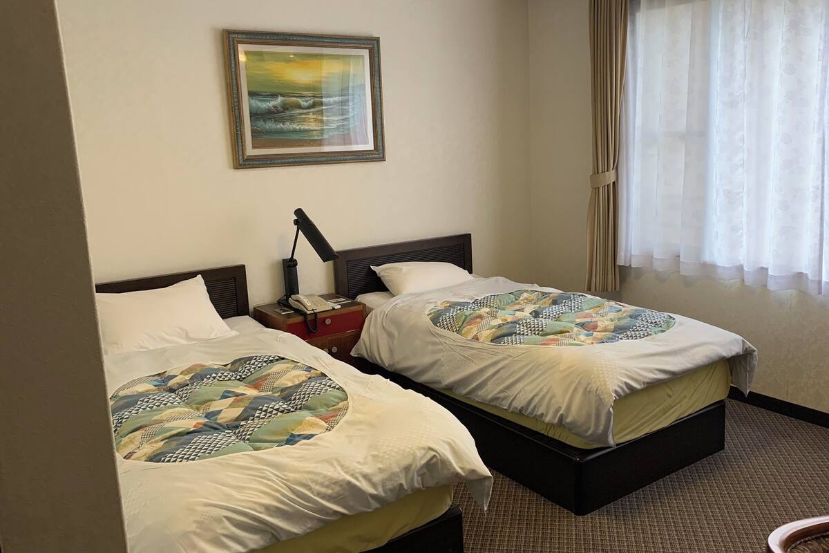 Une chambre d’hôtel avec deux lits, une table de chevet avec une lampe, un tableau au mur et une fenêtre avec des rideaux.