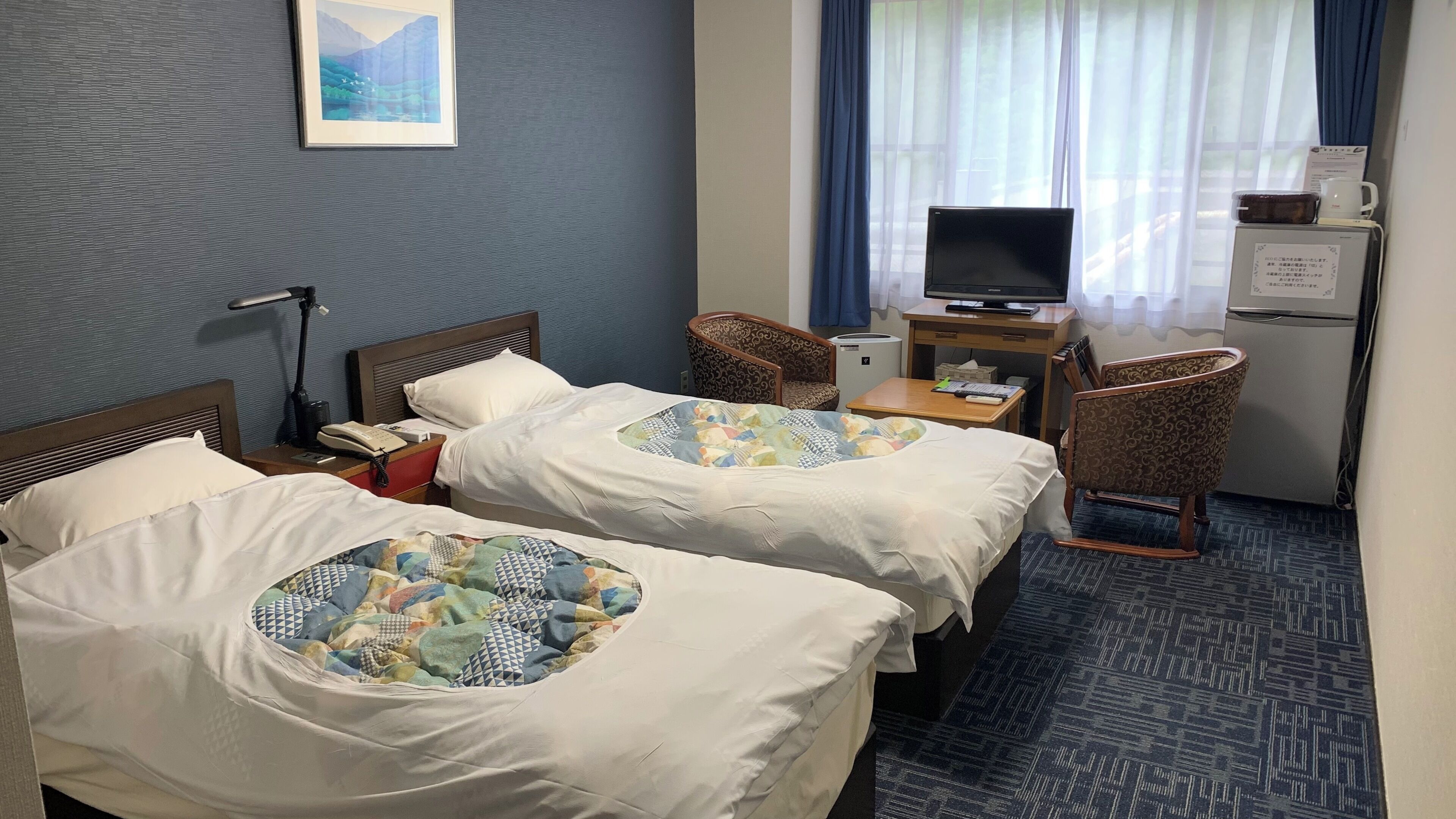 A hotel room with two beds, a desk with a television, a chair, and a small refrigerator.