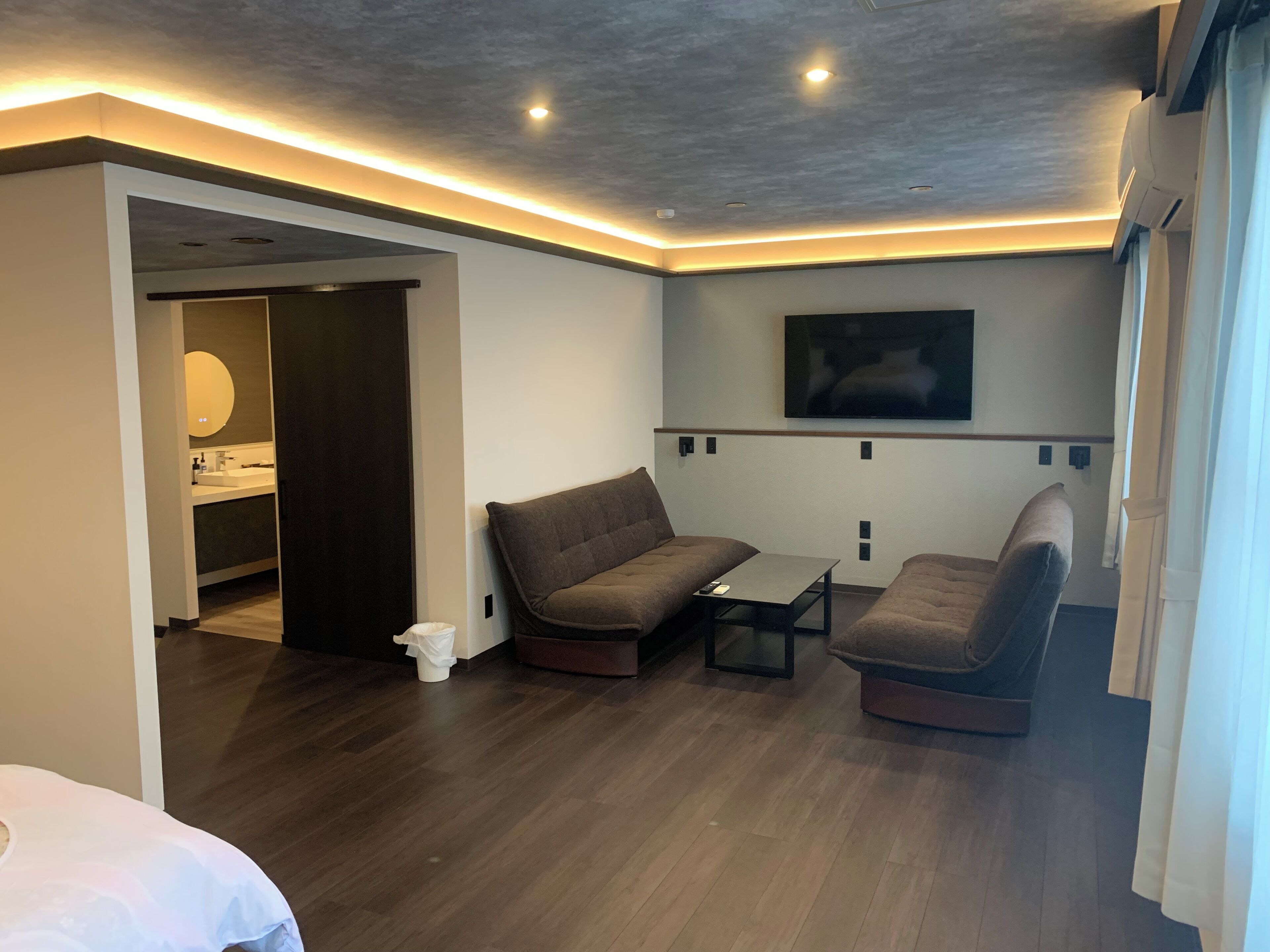 A modern hotel room with a wooden floor, a sofa, a small table, a TV mounted on the wall, and a bathroom visible through an open door.