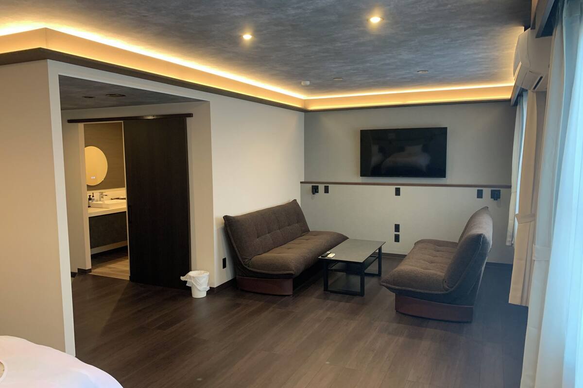Une chambre d’hôtel moderne avec un plancher en bois, un canapé, une petite table, un téléviseur fixé au mur et une salle de bain visible à travers une porte ouverte.