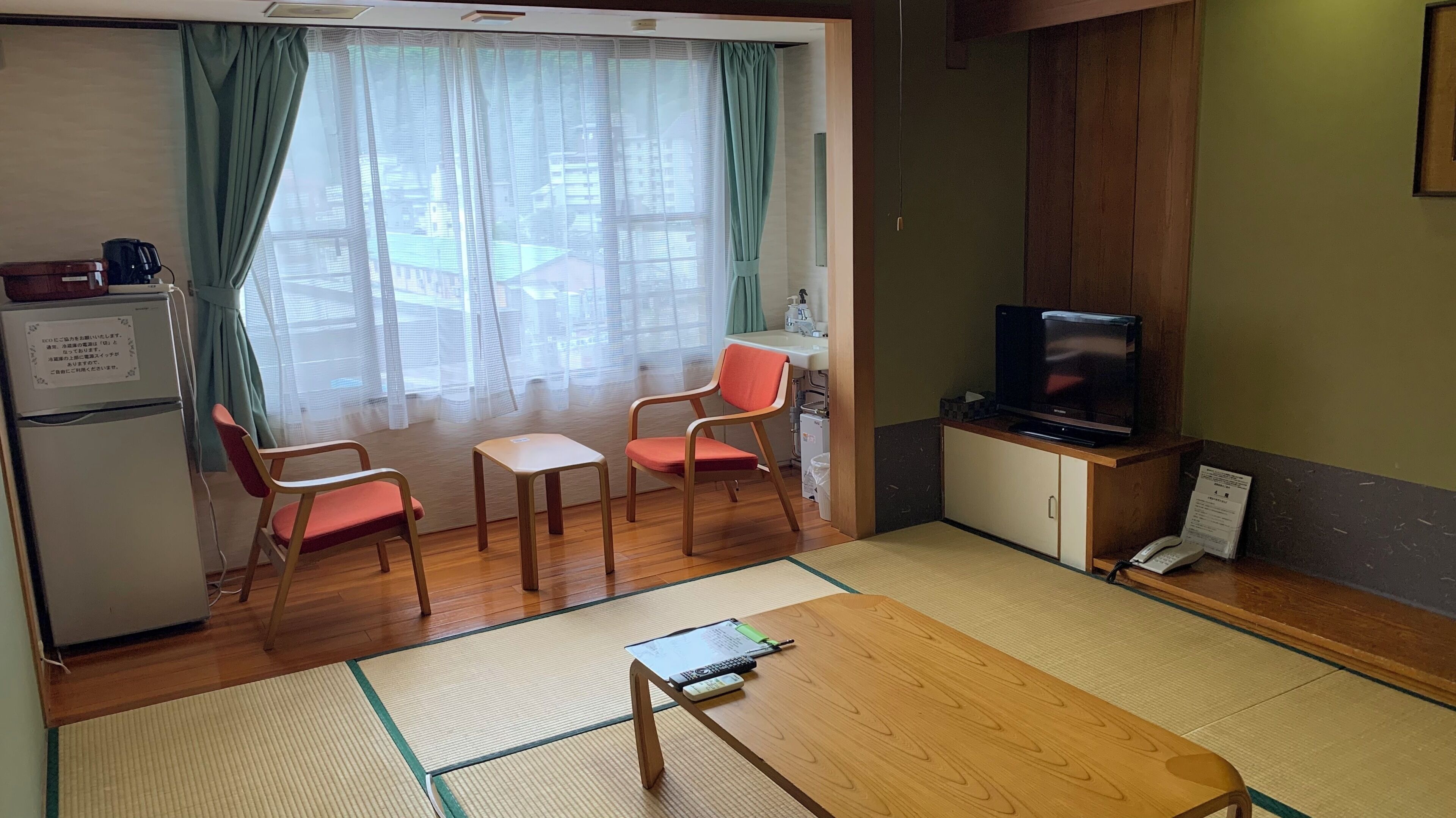 A room with a wooden table, two chairs, a television, a refrigerator, and a window with curtains.