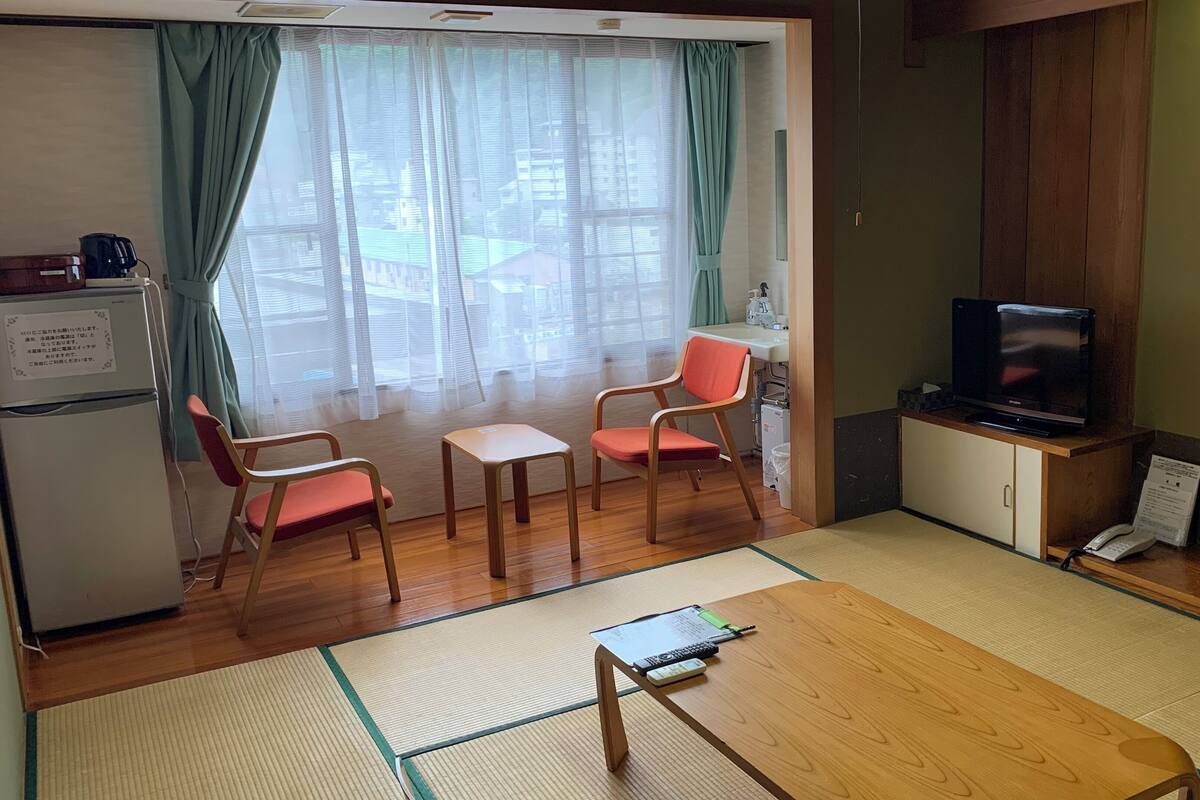 Une pièce avec une table en bois, deux chaises, un téléviseur, un réfrigérateur et une fenêtre avec des rideaux.