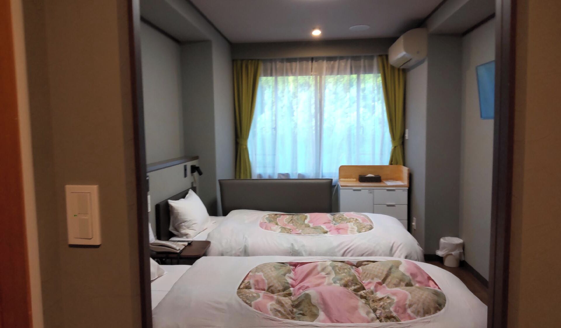 A hotel room with two beds, a desk, a chair, a window with curtains, and an air conditioning unit.