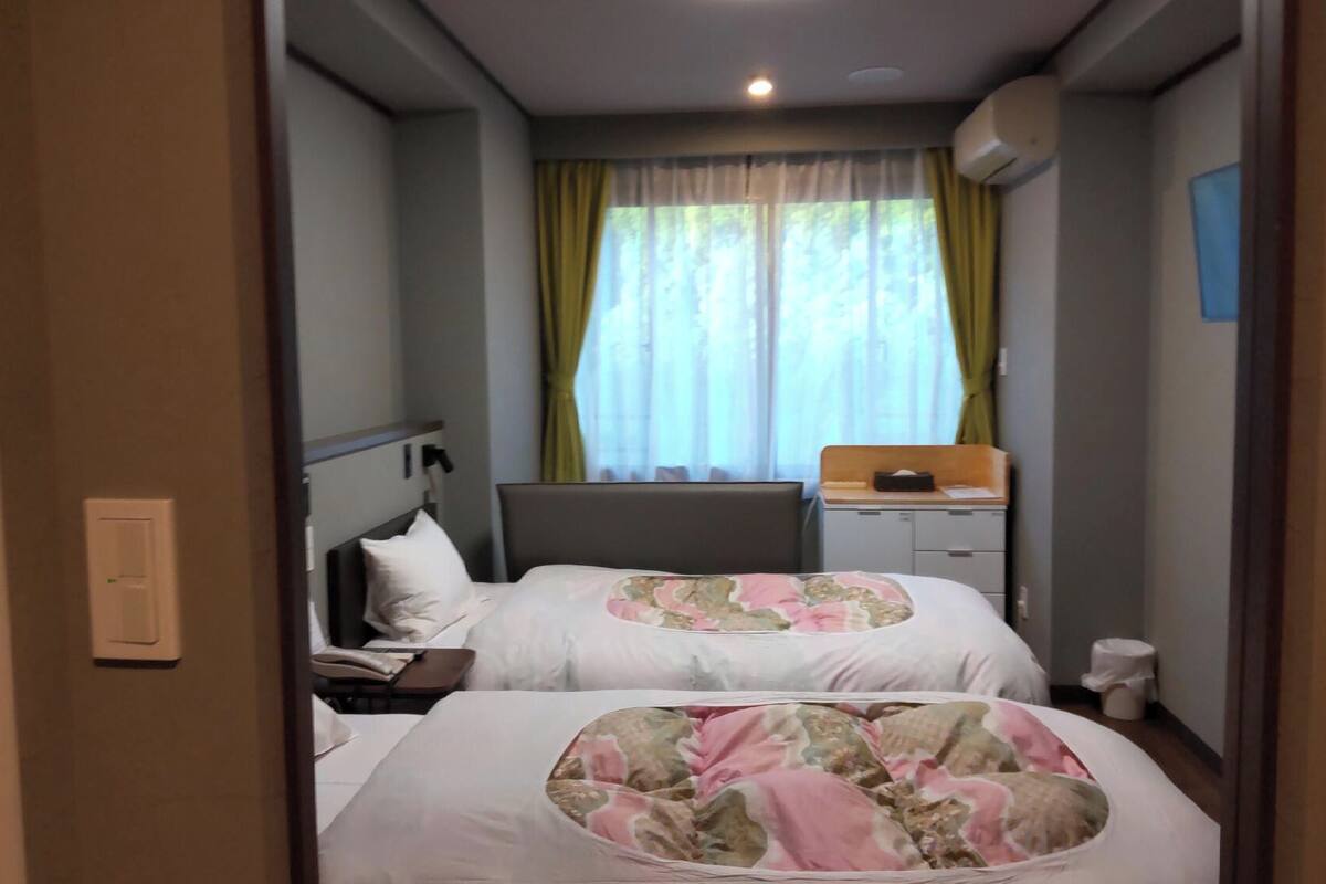 Une chambre d’hôtel avec deux lits, un bureau, une chaise, une fenêtre avec des rideaux et un appareil de climatisation.