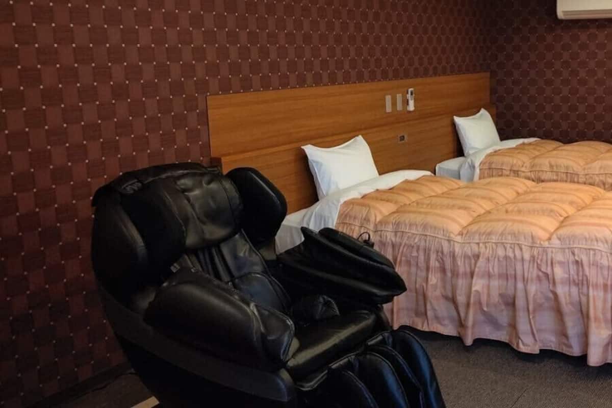 Habitació d'hotel amb una cadira de massatge, dos llits amb roba de llit rosa i un panell de capçal de fusta.