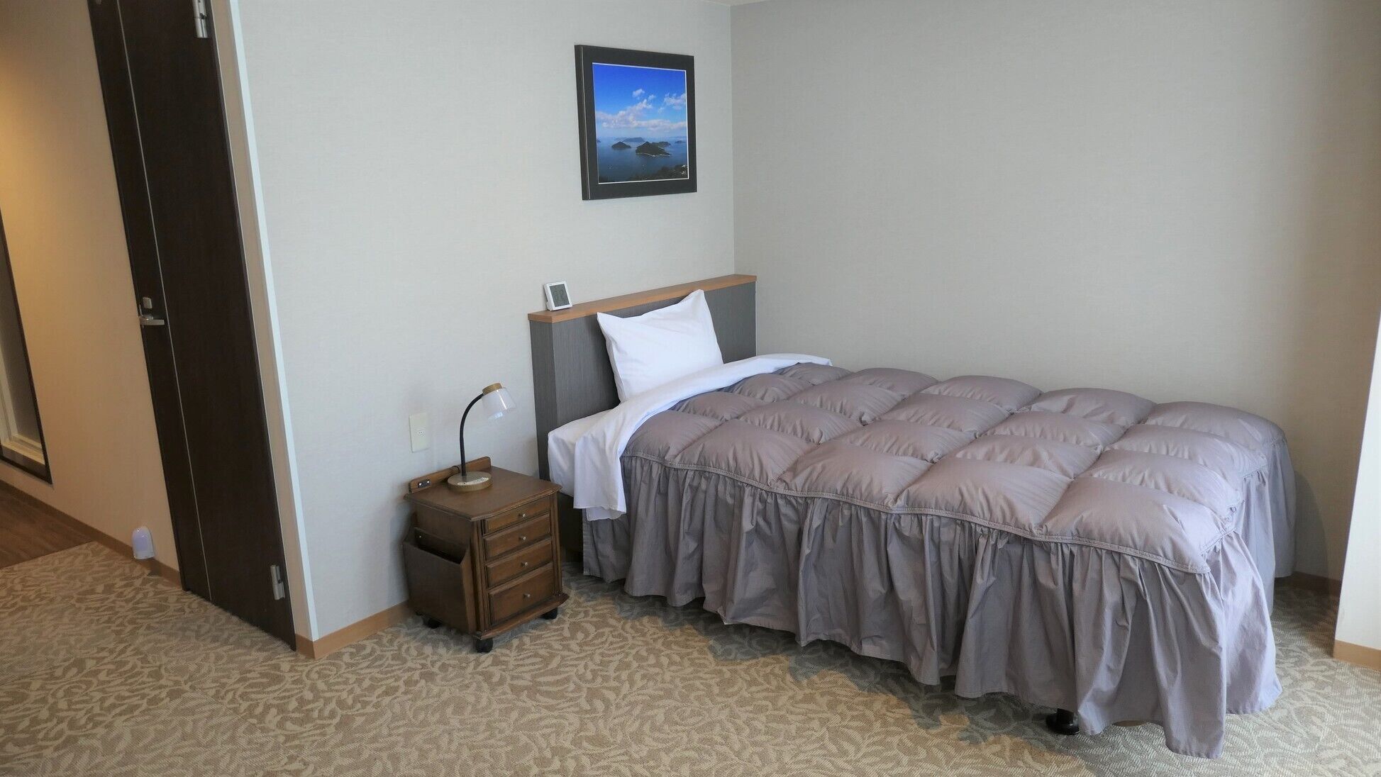 A bedroom with a bed, a nightstand, a lamp, a framed picture on the wall, and a patterned carpet.