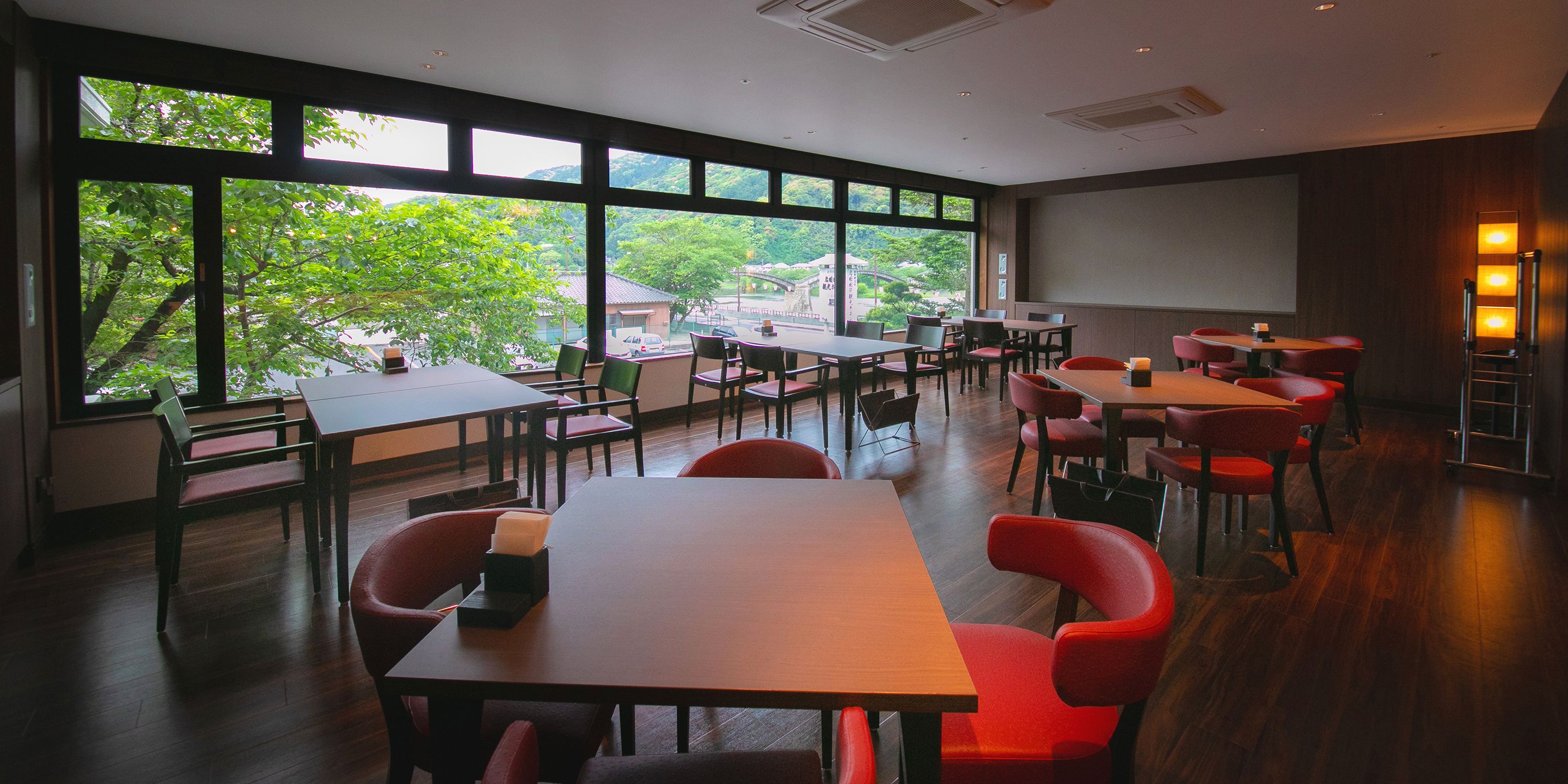 A spacious dining area with multiple tables and chairs, large windows offering a view of greenery and buildings outside.