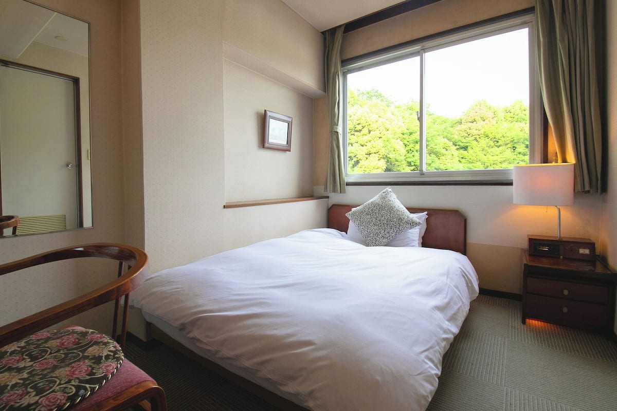 Une chambre d’hôtel avec un lit, une chaise, une table de chevet, une lampe, une fenêtre donnant sur des arbres et un miroir au mur.