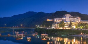 Een hotel aan de rivier met meerdere boten afgemeerd, omringd door weelderige begroeiing en een berg op de achtergrond.