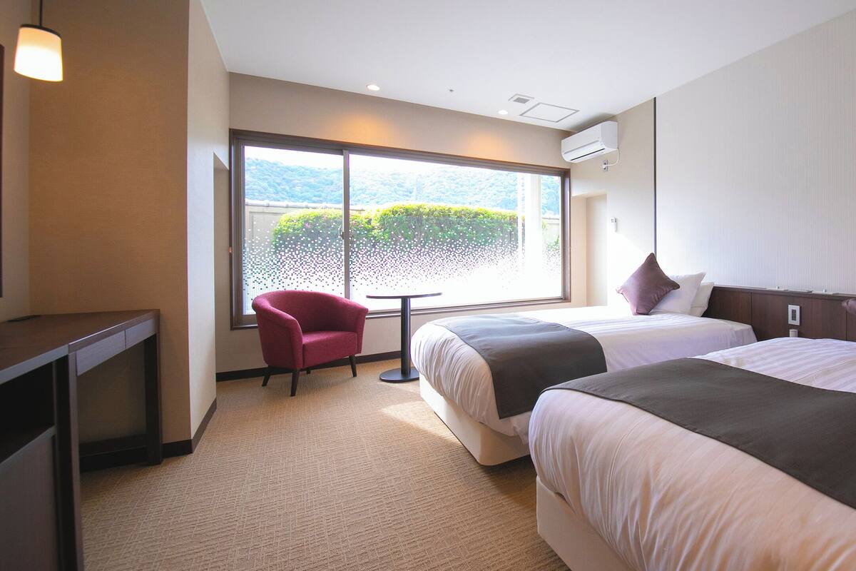 Une chambre d’hôtel avec deux lits, un bureau, une chaise et une grande fenêtre donnant sur un beau paysage.
