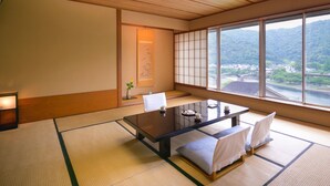 Une pièce de style japonais traditionnel, avec un sol recouvert de tatamis, une petite table basse entourée de coussins et une grande fenêtre donnant sur un pont et la verdure environnante.