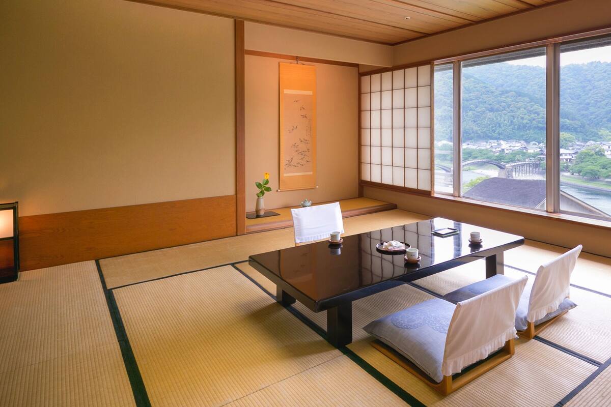 Une pièce de style japonais traditionnel, avec un sol recouvert de tatamis, une petite table basse entourée de coussins et une grande fenêtre donnant sur un pont et la verdure environnante.