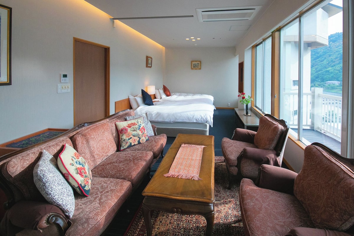 Une chambre d’hôtel avec un canapé, un fauteuil et un lit. Il y a une fenêtre donnant sur un pont et sur la verdure.