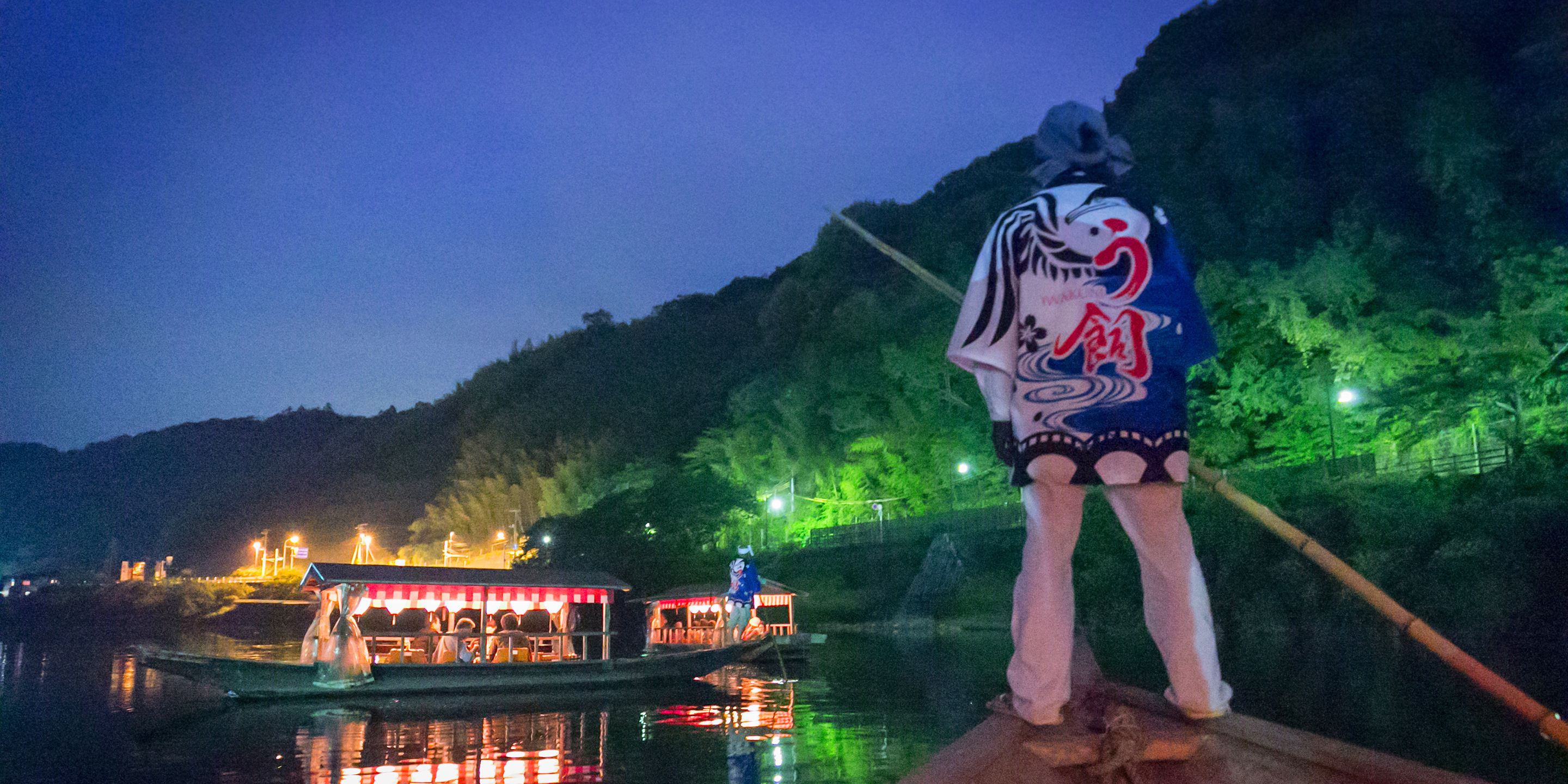 A person standing on a dock by a river at night, with a boat and illuminated buildings in the background.