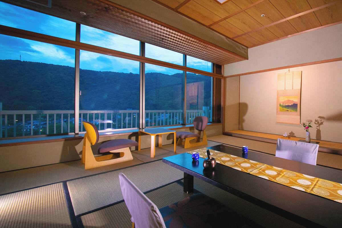 Une pièce avec des tatamis, une table basse agrémentée d’un chemin de table décoratif, des chaises, une petite table avec une tasse bleue, un vase de fleurs et un tableau au mur.