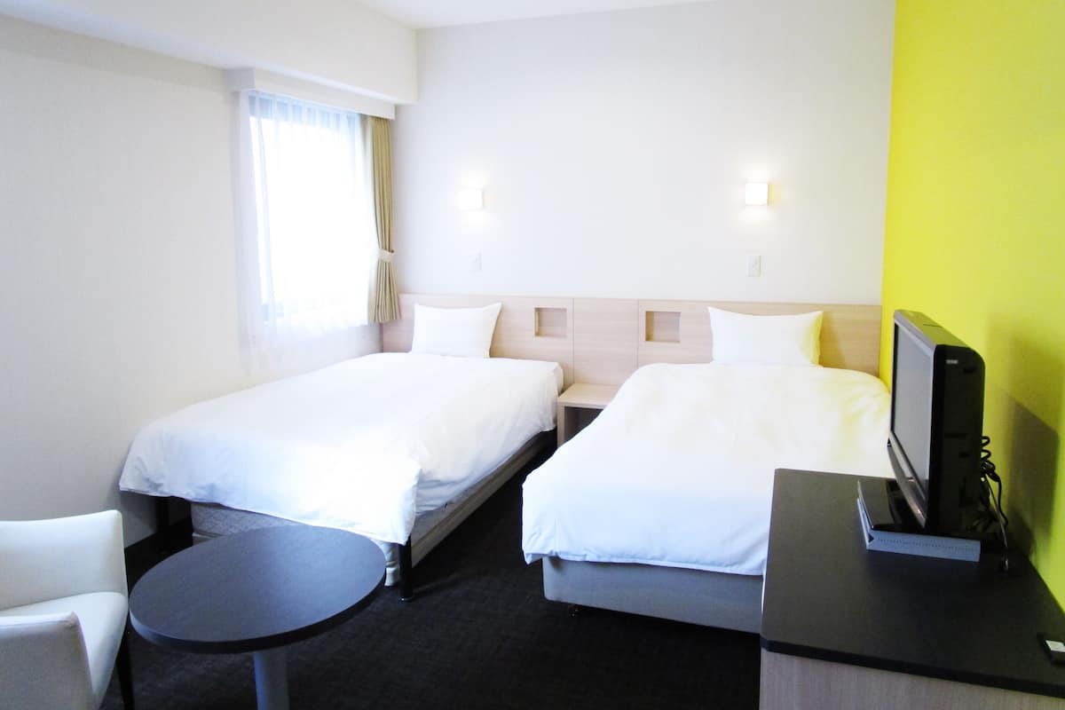 Quarto de hotel com duas camas de solteiro, uma mesinha redonda, uma escrivaninha com computador e uma janela com cortinas.