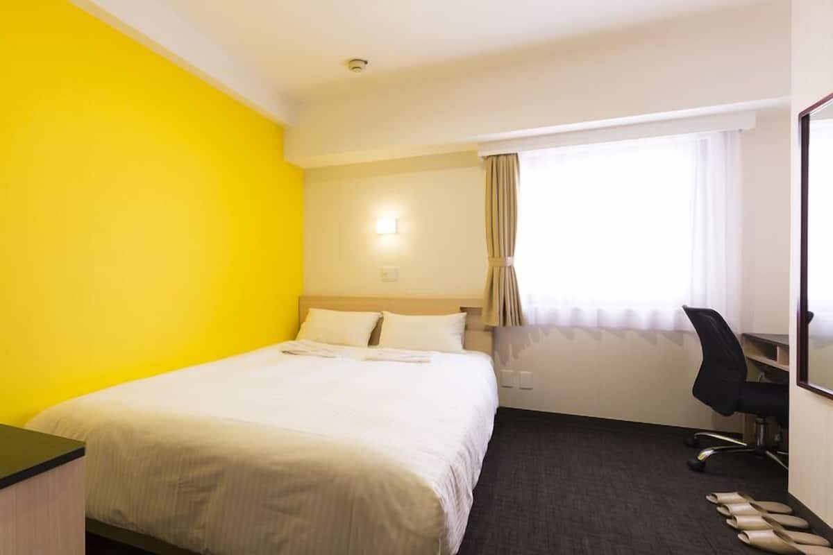 Quarto de hotel com cama, secretária, cadeira e janela com cortinas.