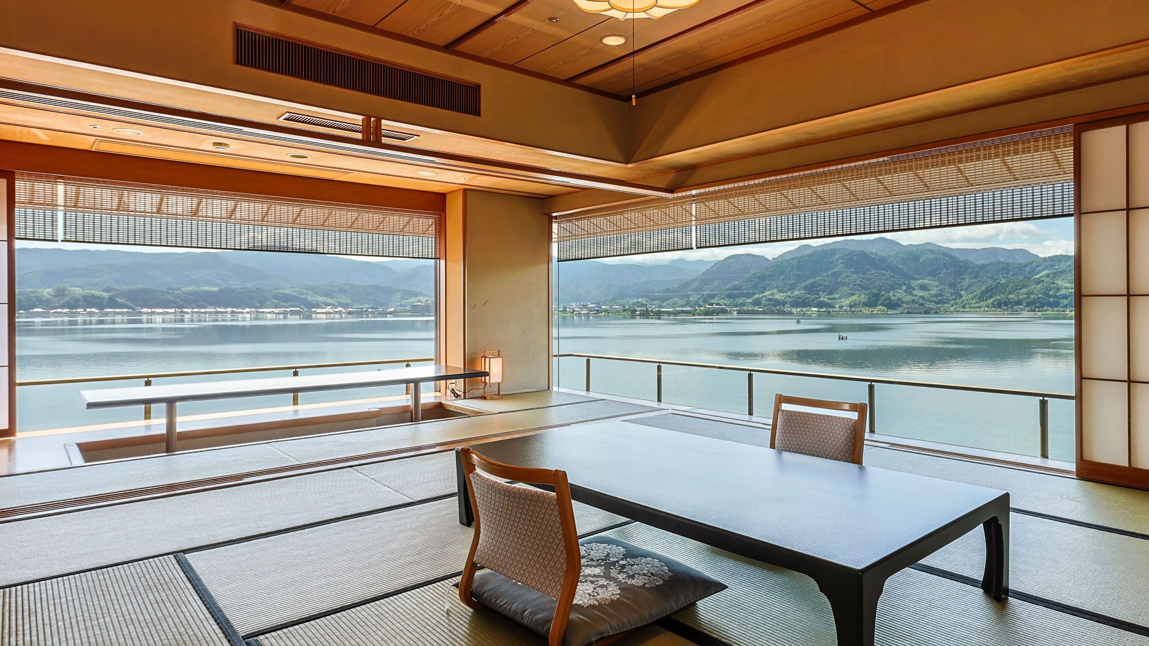 A traditional Japanese-style room with tatami flooring, a low table, and chairs. Large windows offer a view of a lake and mountains.