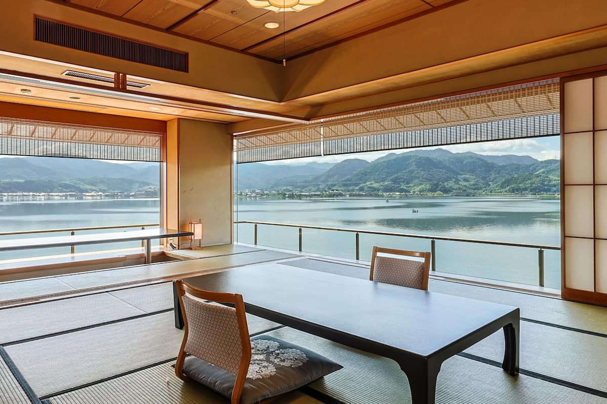 A traditional Japanese-style room with tatami flooring, a low table, and chairs. Large windows offer a view of a lake and mountains.
