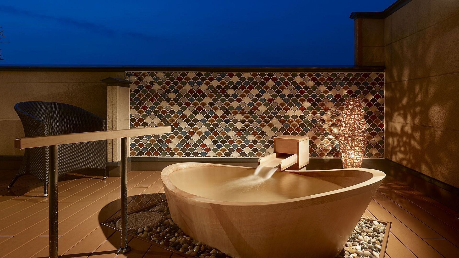 A wooden hot tub with a wooden bench and a chair, set against a tiled wall with a geometric pattern.
